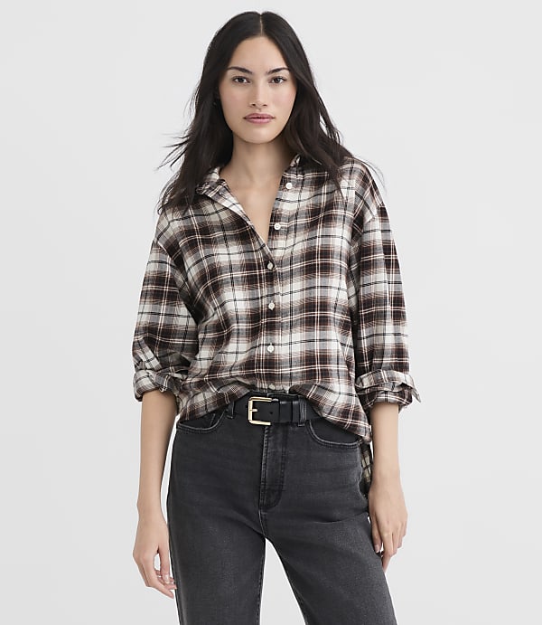 A woman in a brown plaid shirt and black jeans stands against a plain white background.