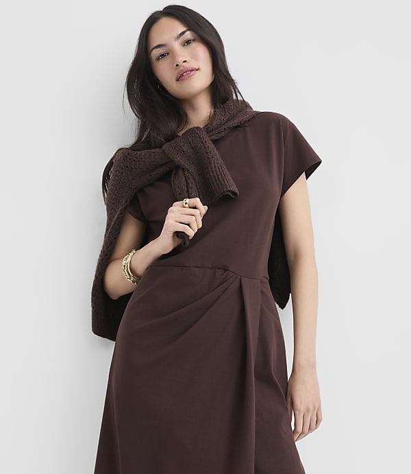 A woman in a brown dress and sweater stands against a plain white background.