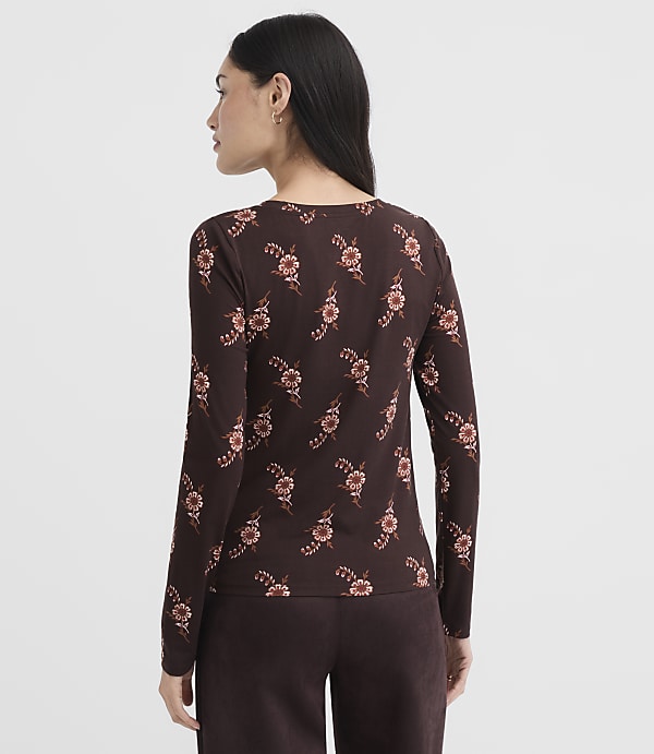 A woman in a brown floral long-sleeved shirt and pants stands against a plain, light brown background.