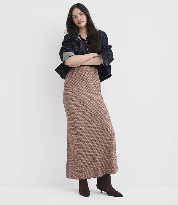 A woman in a patterned skirt and denim jacket stands with her arms crossed against a plain white background.
