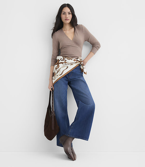 A woman in a brown wrap top and wide-leg jeans stands against a plain white background, holding a brown handbag.