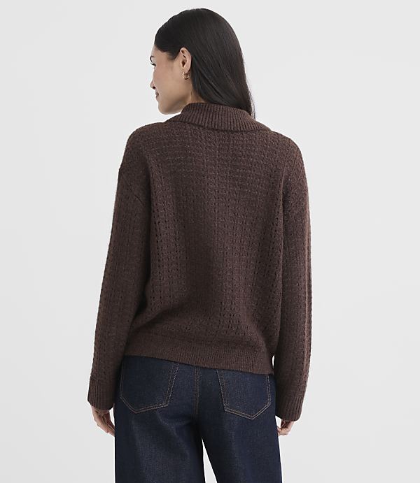 A woman in a brown turtleneck sweater and dark jeans stands against a textured white background.