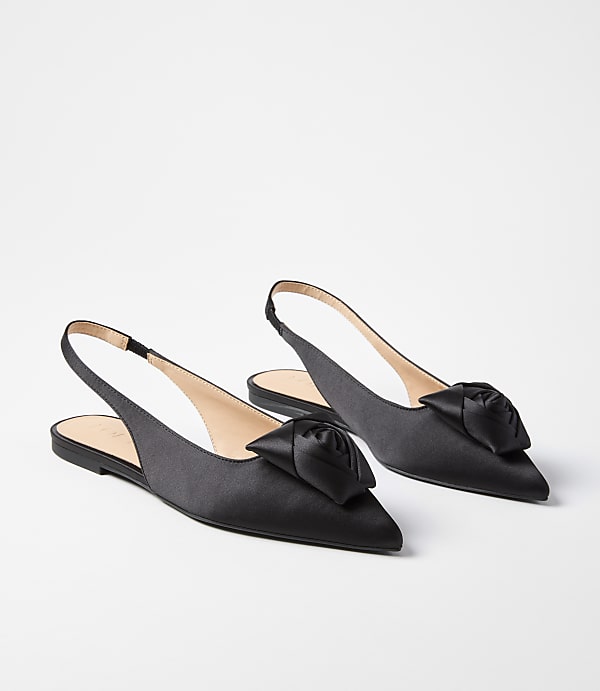 A pair of black satin slingback flats with a rose embellishment on the toe rest on a plain white background.