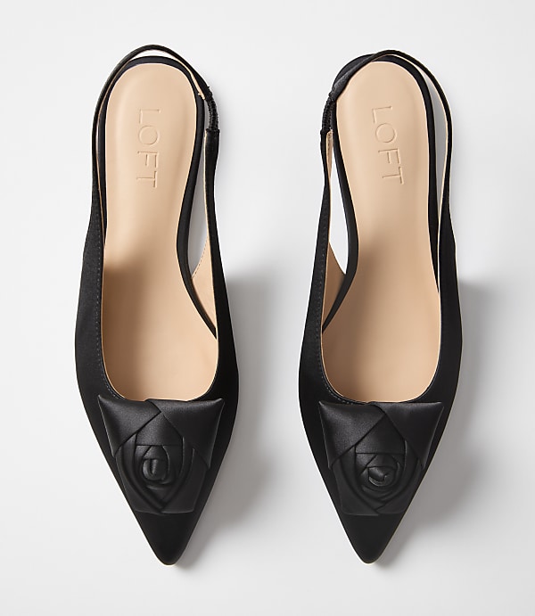 A pair of black satin slingback heels with a rose detail on the toe sit on a plain white background.
