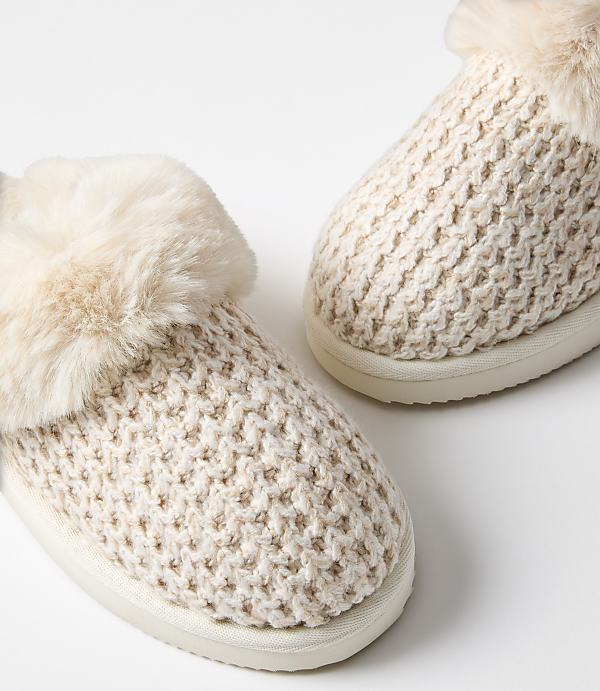 A pair of cozy, cream-colored knitted slippers with fluffy trim rests on a plain white background.