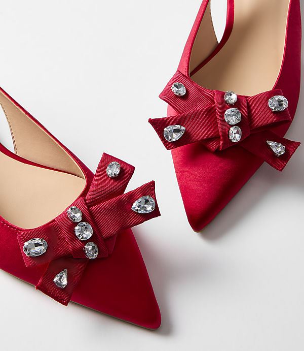 Two red satin pointed-toe heels with jeweled bows are displayed against a plain white background.
