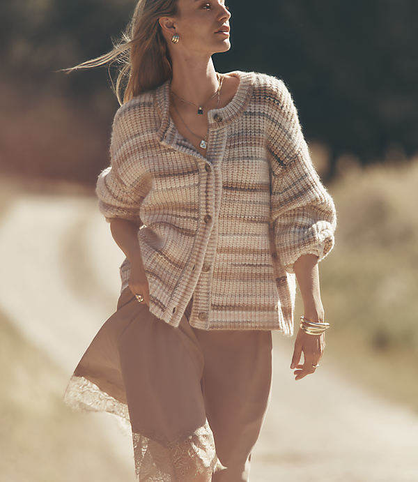 A woman in a striped cardigan and slip dress walks down a dirt road with trees in the background.