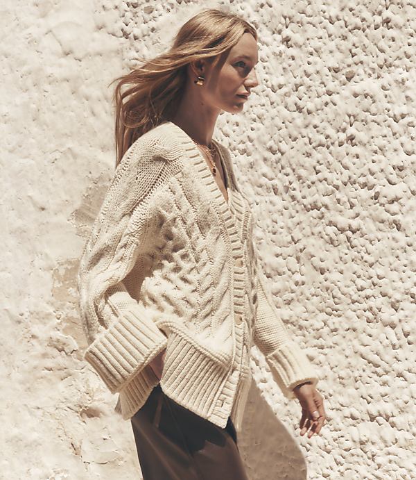 A blonde woman in a cream cable-knit cardigan walks against a textured, light-colored wall.