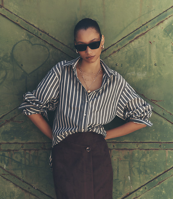 A woman in a striped shirt and dark skirt stands against a green, textured metal door.