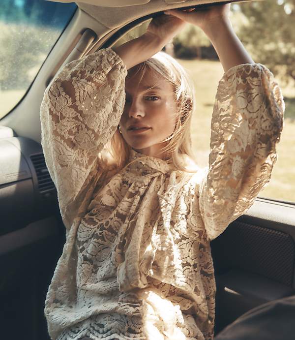 A woman in a lace top leans back in a car, her arms raised to the roof, with a sunlit, grassy landscape visible through the window.