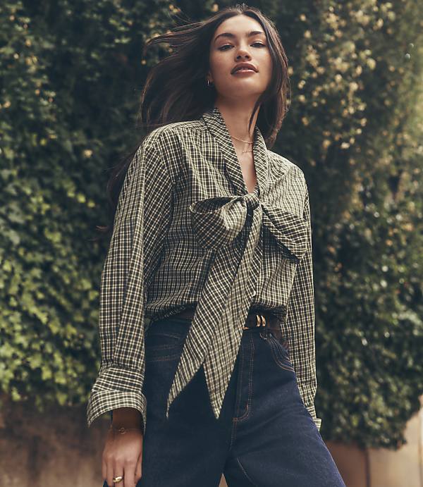 A woman in a checkered blouse with a large bow stands in front of a lush green hedge.
