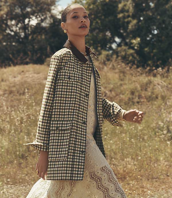 A woman in a checkered jacket and patterned dress walks in a field with trees in the background.