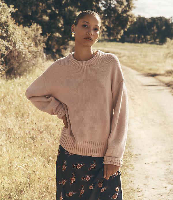 A woman in a pink sweater and floral skirt stands on a dirt path with trees and fields in the background.