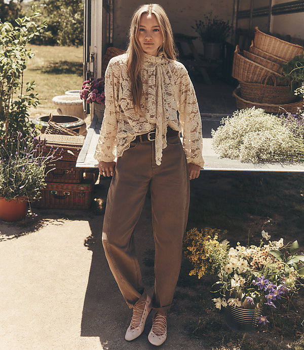 A young woman in a lace top and brown pants stands in front of a truck filled with flowers and baskets, with greenery in the background.