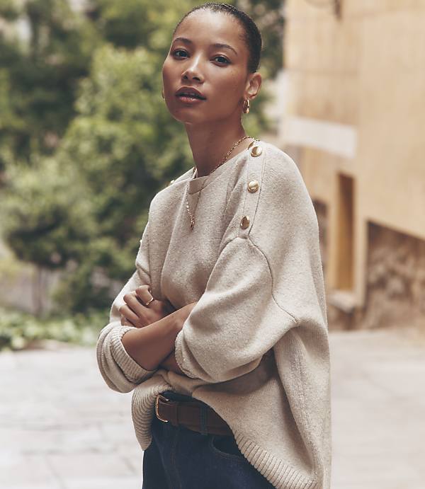 A woman in a cream-colored sweater with gold buttons stands with her arms crossed in front of a blurred background of a European street.