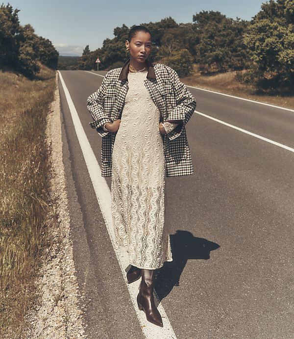 A woman in a patterned jacket and lace dress stands on an empty asphalt road with grassy verges, leading towards a tree-lined horizon under a clear sky.