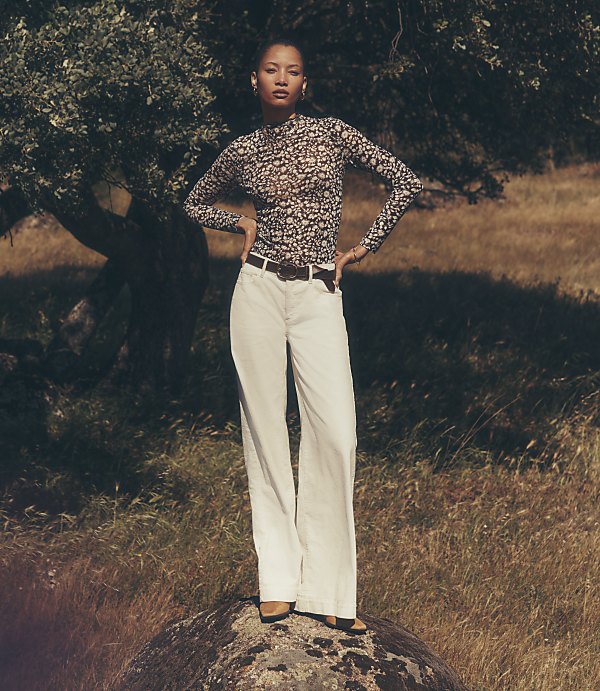 A woman in a patterned top and white pants stands on a rock in a grassy field with a large tree behind her.