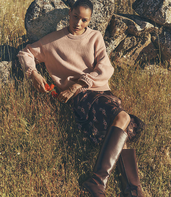 A woman in a pink sweater and floral skirt rests against rocks in a grassy field.