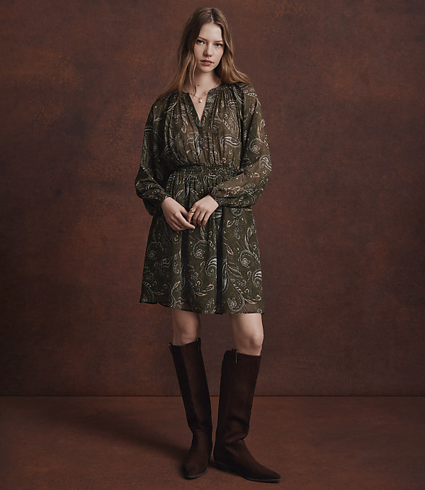 A woman in a patterned green dress and brown boots stands against a textured brown background.