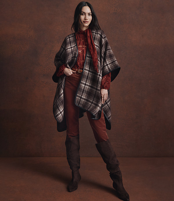 A woman in a plaid poncho and rust-colored pants stands against a textured brown background.