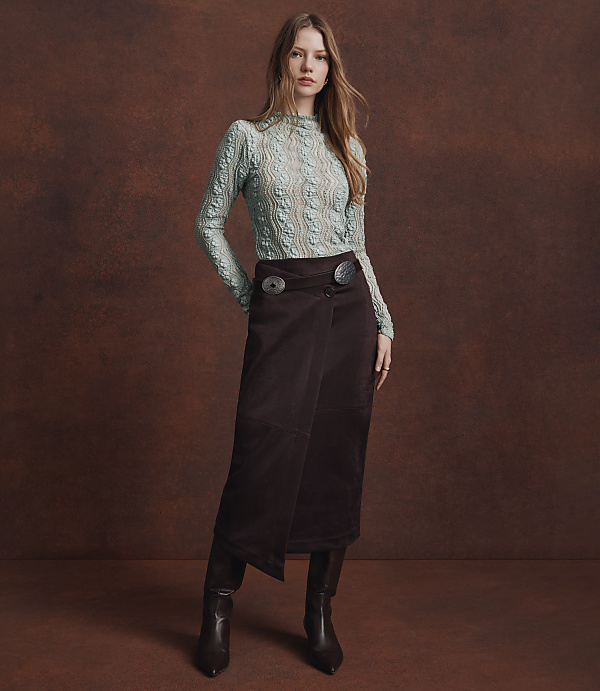 A woman in a light blue textured top and a dark brown wrap skirt stands against a mottled brown background.