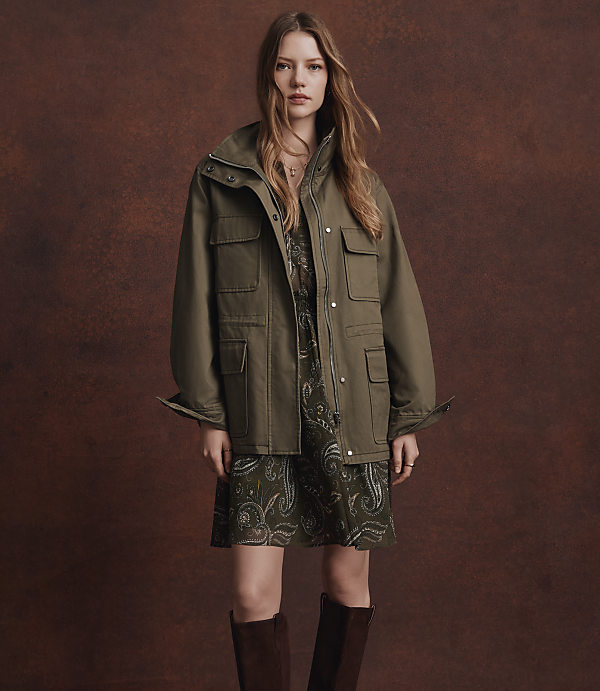 A woman in a green jacket and dress stands against a textured brown background.