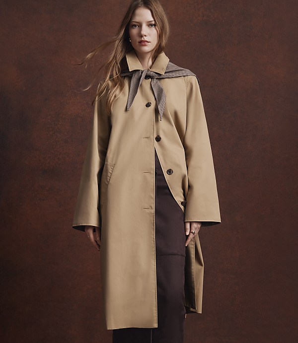 A woman in a tan trench coat stands against a textured brown background.