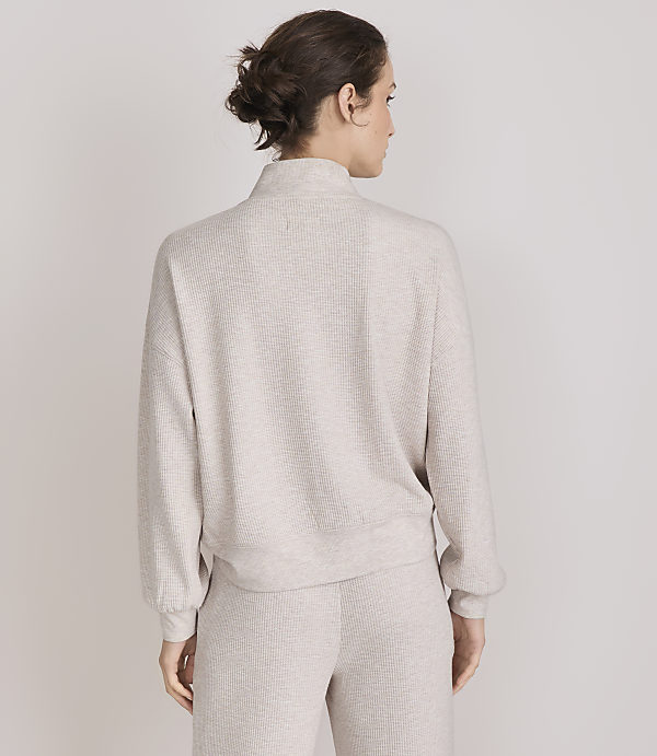 A woman in a beige mock-neck sweater and matching pants stands against a plain white background.