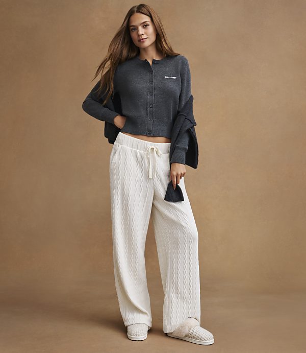 A young woman in a dark gray cardigan and white cable-knit pants stands against a plain brown background.