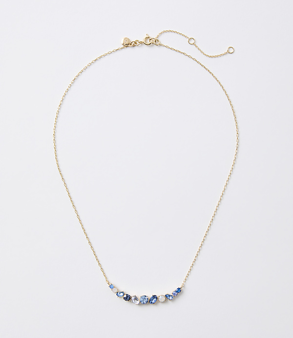 A delicate gold necklace with a curved arrangement of blue and white gemstones rests on a plain white background.