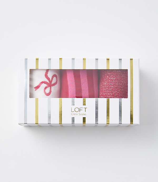 A white box with gold and silver stripes contains three pairs of pink socks, one with a bow, one striped, and one glittery, against a plain white background.