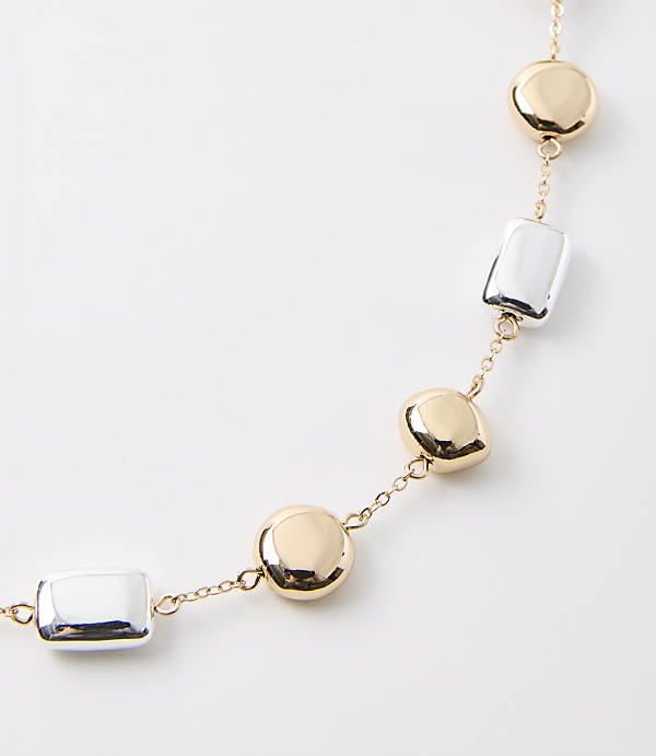 A delicate gold chain necklace with alternating polished gold discs and silver rectangular beads rests on a plain white background.