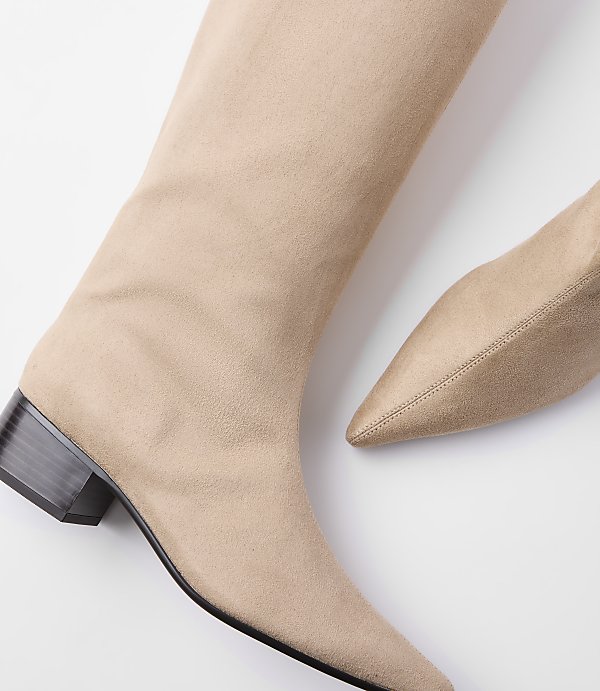A pair of beige suede boots with pointed toes and block heels are laid out on a white background.