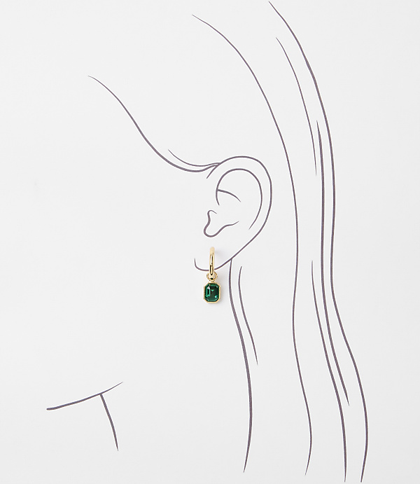 A delicate gold hoop earring with a small emerald gemstone dangles from a stylized outline of an ear against a plain white background.