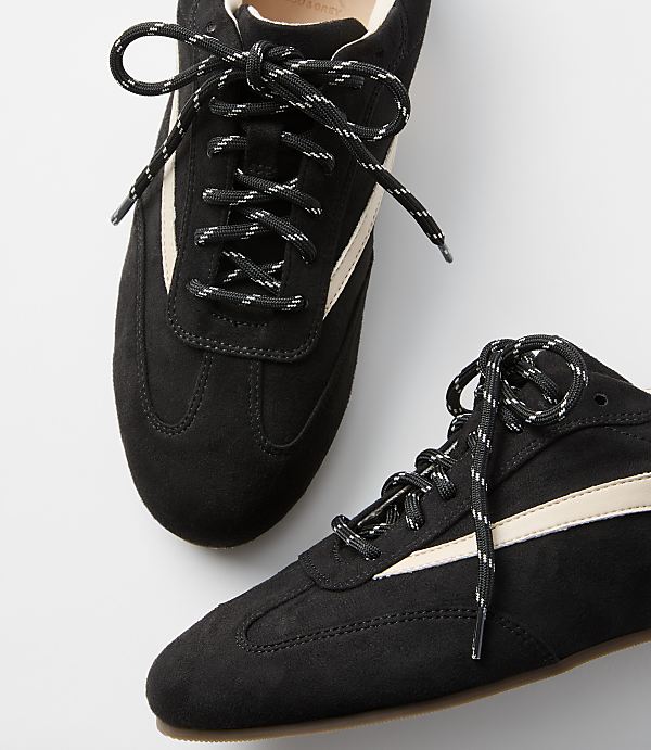 A pair of black suede sneakers with cream stripes and black and white laces rests on a plain white background.