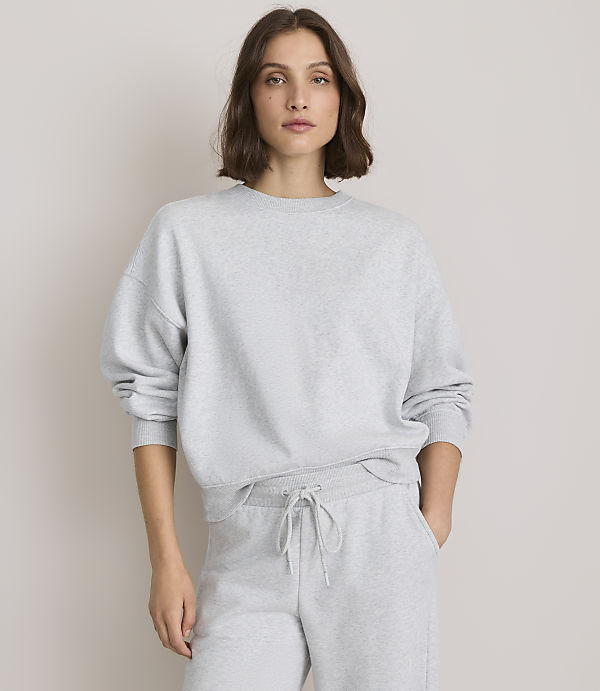 A woman in a grey sweatshirt and sweatpants stands against a plain, light grey background.