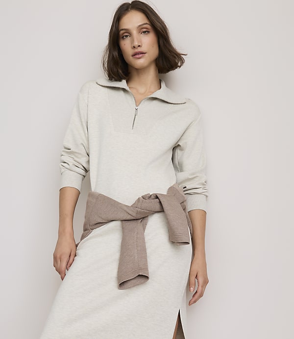 A woman in a light gray collared dress with a brown sweater tied around her waist stands against a plain, light-colored background.