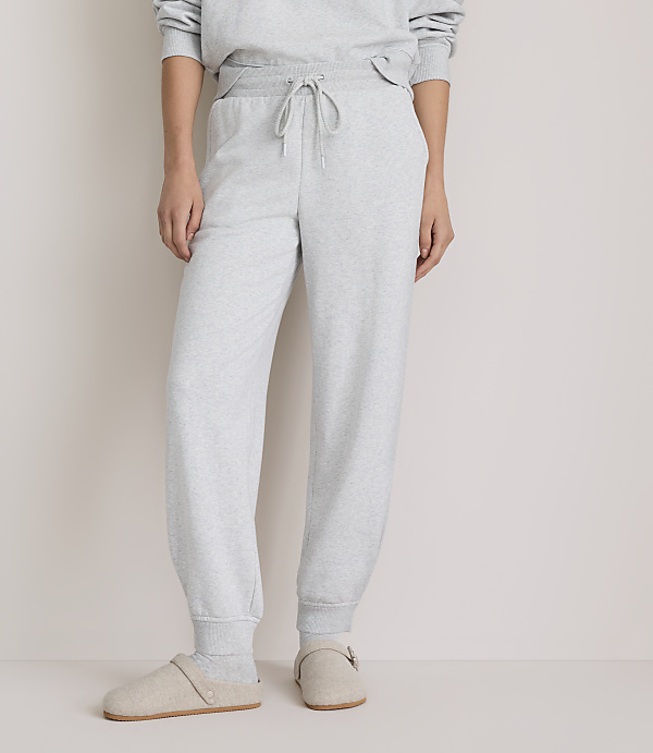 A person wearing light grey sweatpants and slippers stands against a plain, light grey background.