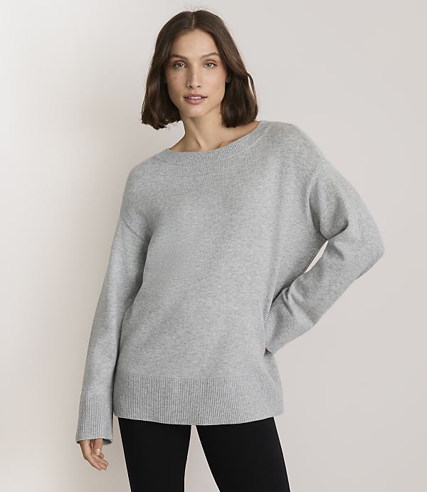A woman in a light gray sweater and black leggings stands against a plain white background.