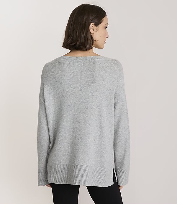 A woman in a grey sweater stands against a plain, light grey background.