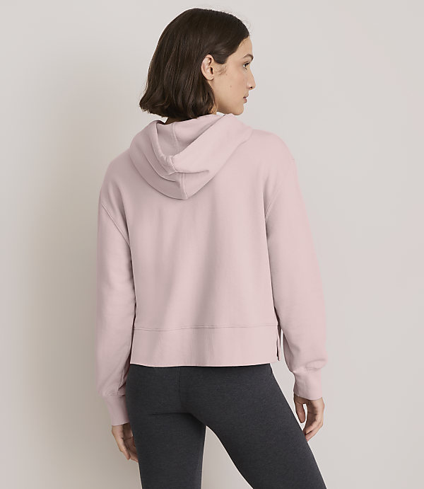 A woman in a pink hoodie and dark leggings stands against a plain, light gray background.