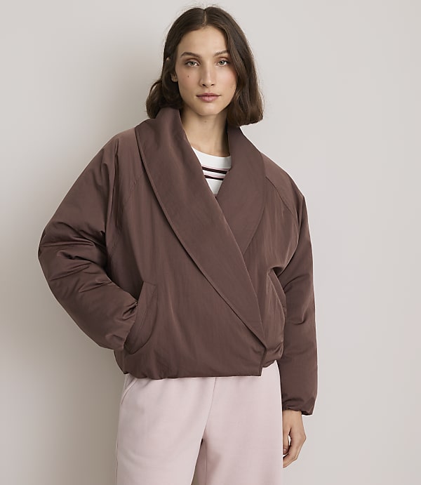 A woman in a brown puffer jacket and pink pants stands against a plain, light-colored background.