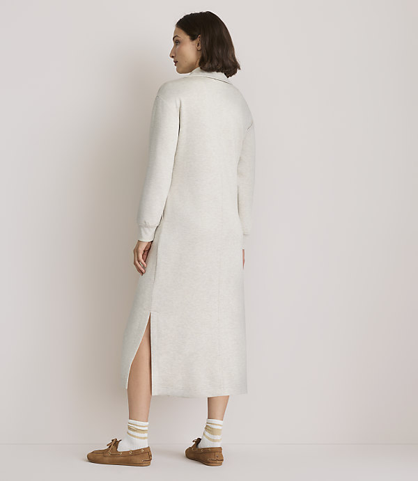 A woman in a light-colored sweater dress and loafers stands against a plain, light-colored background.