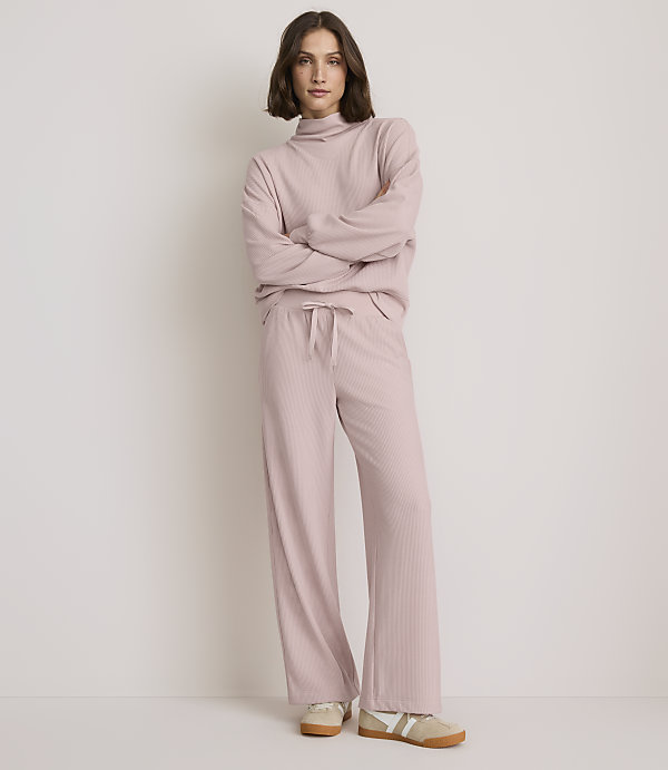 A woman in a pink ribbed loungewear set stands with her arms crossed against a plain, light-colored background.