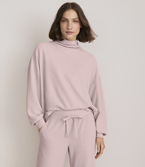 A woman in a pink ribbed turtleneck sweater and matching pants stands against a plain white background.