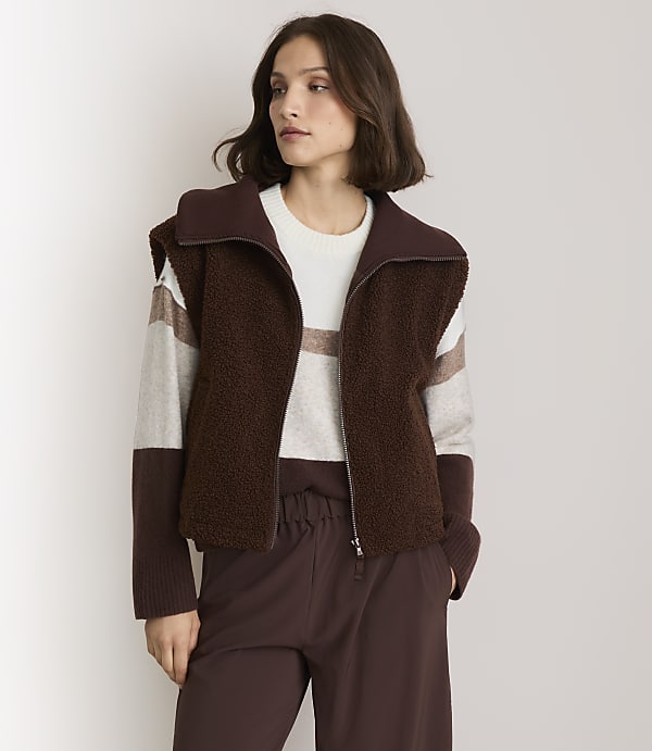 A woman in a brown fuzzy vest and striped sweater stands against a plain white background.