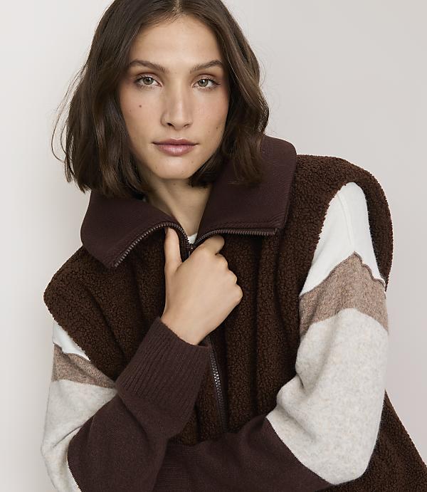 A woman in a brown fleece vest and striped sweater poses against a plain background.