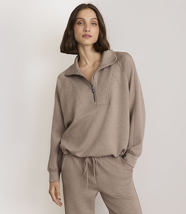 A woman in a taupe quarter-zip sweatshirt and matching sweatpants stands against a plain, light gray background.