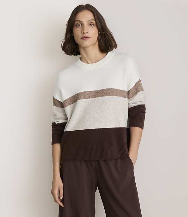 A woman in a striped sweater and brown pants stands against a plain white background.