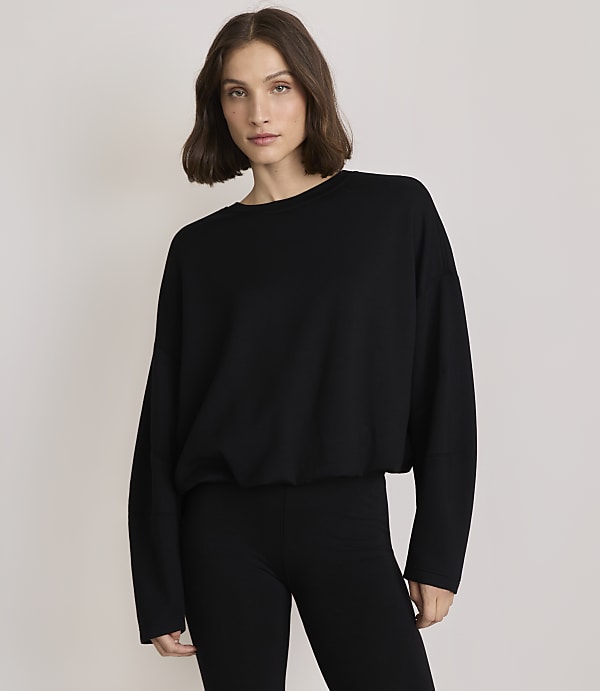 A woman in a black sweater and leggings stands against a plain, light gray background.